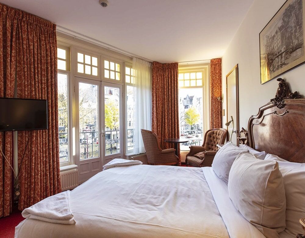 Double Room with Canal View: a carved wooden bed facing tall canal-side windows hung with red drapes